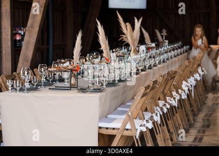 A long wedding reception table is beautifully set inside a rustic barn, featuring beige linens, wooden chairs with white ribbons, and pampas grass cen Stock Photo