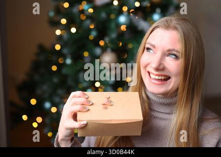 Blonde woman holding gift box with amazed expression, cozy winter holiday atmosphere Stock Photo