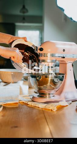 A person is making a cake in a pink mixer. The mixer is on a wooden table. The cake batter is poured into the mixer Stock Photo