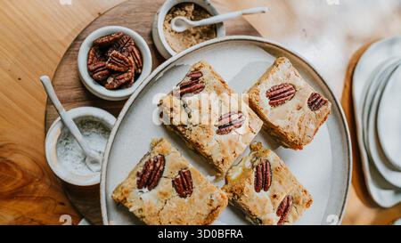 A plate of four peanut butter bars sits on a wooden table. The bars are topped with nuts and are surrounded by small bowls containing sugar and salt. Stock Photo