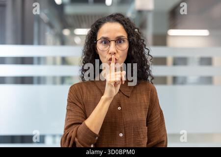 Woman with curly hair and glasses standing in an office, holding a finger to her lips, making a shushing gesture representing secrecy, quiet, and privacy Stock Photo