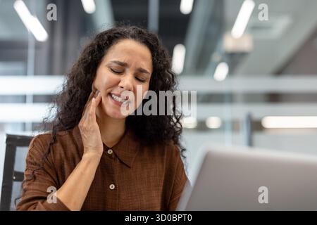 Woman suffering from a painful toothache, holding her jaw and closing her eyes in discomfort, struggling to concentrate on working with her laptop at her office desk Stock Photo