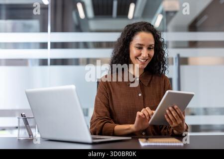 Professional businesswoman with curly hair smiling while using a tablet at a modern desk, multitasking with laptop, showcasing confident remote work, digital connectivity and success Stock Photo