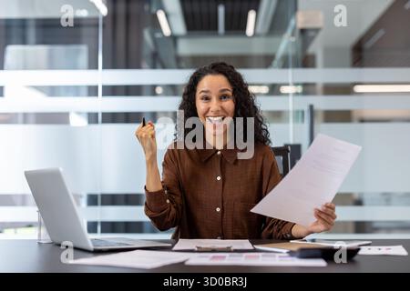 Young mixed race businesswoman celebrating achievement and feeling excited, holding success document and gesturing with a joyful expression while working in a bright office environment Stock Photo