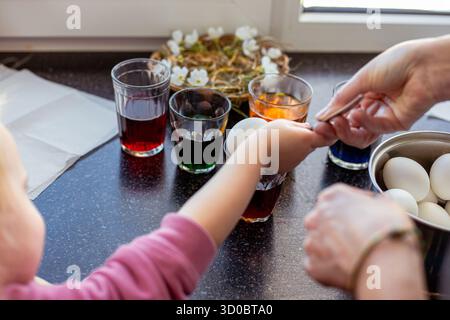 A mother and child paint Easter eggs together in different colors at a table by the window. Stock Photo