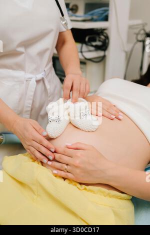 Pregnant woman and gynecologist with pair of knitted baby booties on her tummy. Expecting mother with tiny baby booties while waiting for her baby Stock Photo