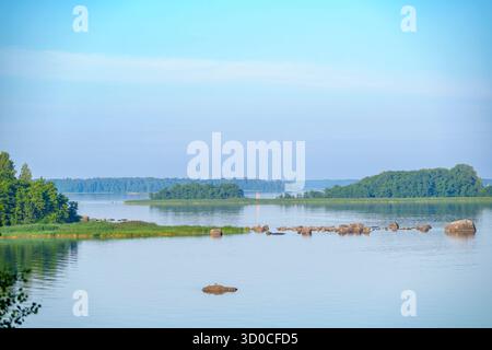 Calm lake scene with scattered rocks, grassy shore, and small forested islands in soft evening light under a pastel-colored sky Stock Photo