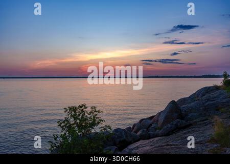 Peaceful sunset view over a body of water with rocks and vegetation in the foreground and soft orange and blue tones in the sky Stock Photo