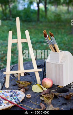 Artistic autumn still life with painting supplies: easel, brushes, palette, apple, fallen leaves on a tree stump in a park. Stock Photo