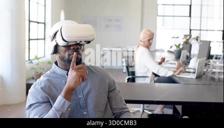 Making shushing gesture Black man wearing VR headset at shared office desk, with laptop coffee mug Stock Photo