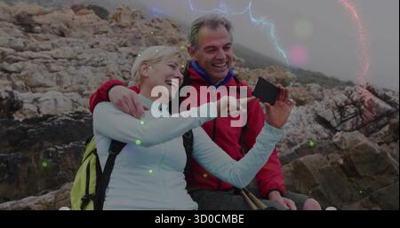 Posing couple wearing outdoor gear on rocky coastal outcrop, holding smartphone and hiking poles Stock Photo