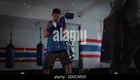 Practicing boxer wearing blue top and gloves throwing punches in gym, with trainer holding pad Stock Photo