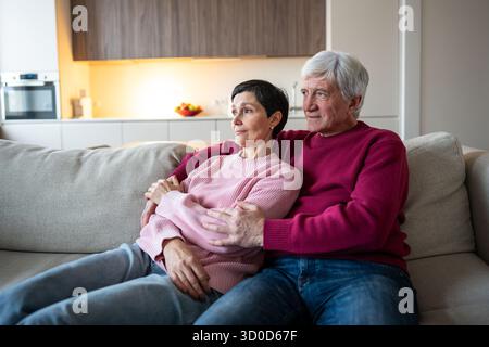 Content elderly couple lies embraced on sofa watching television in calm living room, senior love Stock Photo