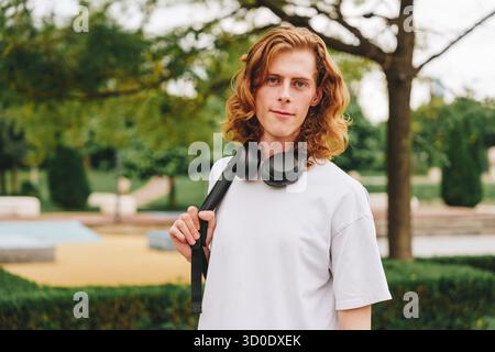 Young man with curly hair stands confidently in a park holding headphones Stock Photo