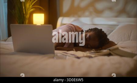Tired exhausted African American ethnic woman female lady girl student lying bed sleeping opened laptop computer working insomnia studying homework Stock Photo