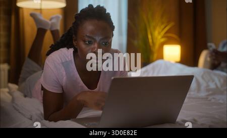 Family weekend relaxed calm African American woman girlfriend browsing internet typing laptop computer lying on bed man boyfriend chatting mobile Stock Photo