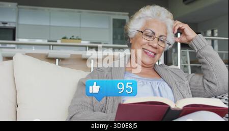 Smiling senior woman in grey cardigan and eyeglasses reading red book on sofa, with modern kitchen Stock Photo
