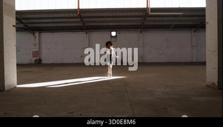 Balancing ballet dancer holding en pointe in abandoned warehouse, with sunlight beam, steel columns Stock Photo
