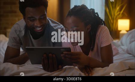 Lovely laughing African American family married couple woman wife man husband lying on bed using digital tablet watch film series online shopping Stock Photo