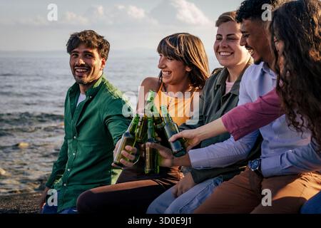 A happy multi-ethnic group of five young friends are laughing and making a toast with green beer bottles while sitting by the sea during a summer even Stock Photo