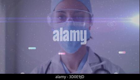 Standing doctor wearing mask cap goggles and coat in clinic, with stethoscope and digital overlay Stock Photo