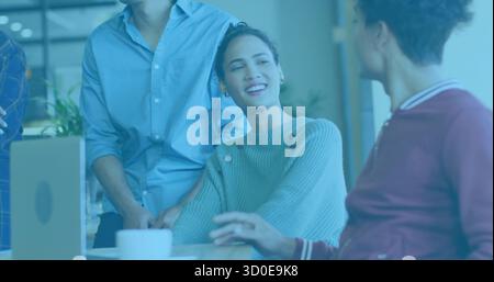 Smiling mid adult Hispanic woman speaking to colleagues at office desk, with laptop and coffee mug Stock Photo