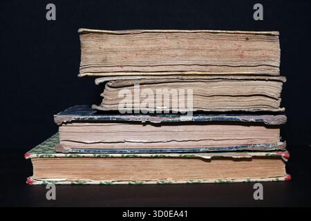 Stack of four heavy antique leather-bound books on a black background, showing the texture of worn, aged pages, spines, and tattered covers. Stock Photo