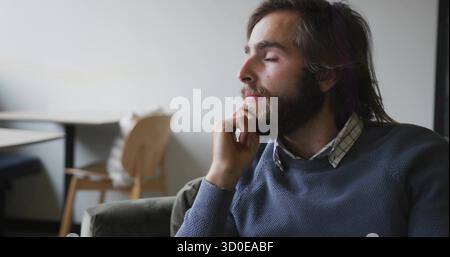 Sitting bearded man resting chin on hand in living area, with cushioned armchair, copy space Stock Photo