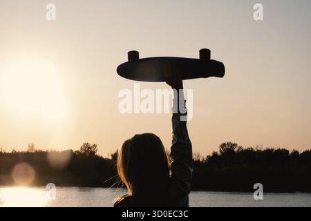 Silhouetted Woman posing with her hands holding skateboard raised up in the sunset sky, summer farewell concept Stock Photo