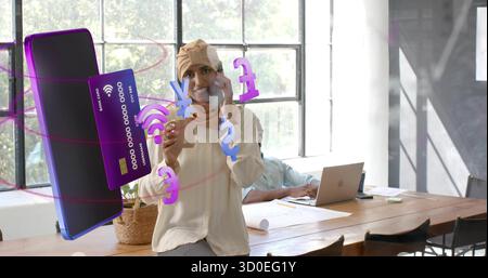 Speaking on smartphone, woman wearing headscarf at office table with floating 3D finance icons Stock Photo