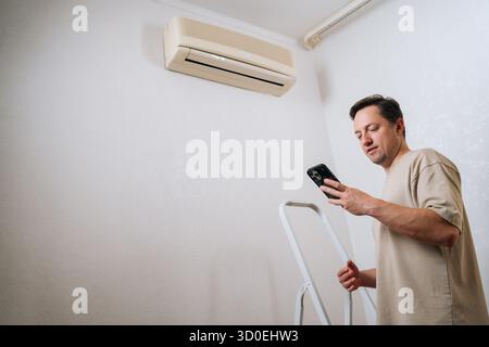Shooting from below of technician male standing by ladder, using smartphone app to control and prepare for air conditioner maintenance, ensuring Stock Photo