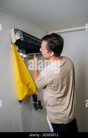 Technician disassembling and repairing wall-mounted air conditioner unit, using drainage bag to collect water while working on electrical components. Stock Photo