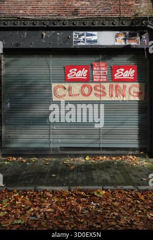 Closed shop with metal shutters, red “Sale” and “Closing Down” signs, fallen autumn leaves on pavement, symbolizing retail decline and closure. Stock Photo