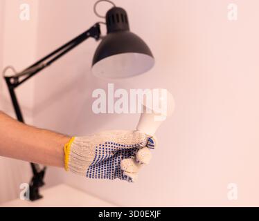 Close-up of a hand in protective glove performing electrical maintenance and installation at home. Electrician replacing a light bulb in a ceiling fix Stock Photo