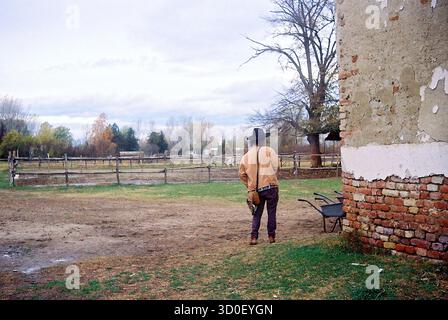 Long-haired man wearing a hat standing beside a weathered ranch house in autumn, captured on 35mm analog film with a timeless rural atmosphere. Stock Photo