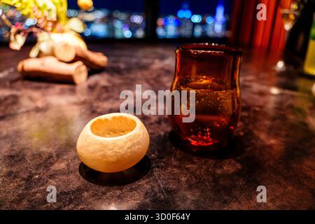 An empty, decorative carved shell bowl and an amber glass with a sparkling beverage sit on a dark table overlooking a city at night. Stock Photo