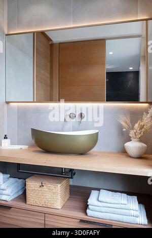 Modern and clean bathroom vanity featuring a stylish countertop washbasin on a wide wooden shelf, a large frameless mirror across the wall Stock Photo