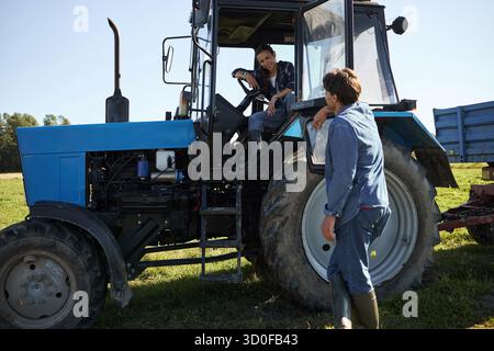 Caucasian young woman sitting in tractor cab talking to Caucasian young man standing beside vehicle on farm field, both smiling and engaging in friendly conversation outdoors Stock Photo