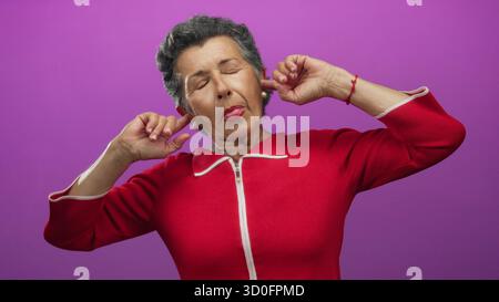 Senior woman with grey hair wearing red blouse standing in front of a vibrant pink background covering her ears with hands in different poses. Stock Photo