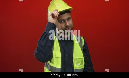Man tipping yellow hardhat with hand while wearing high visibility vest in red studio; confidence safety reliability. Stock Photo