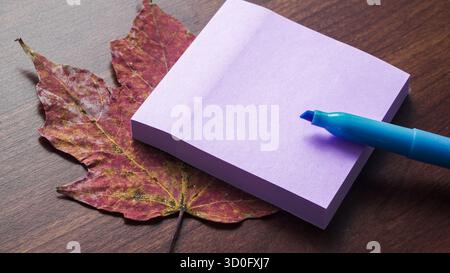 A purple sticky note is placed on a vibrant red maple leaf with a blue marker next to it, all set on a warm wooden table. This scene captures a simple Stock Photo