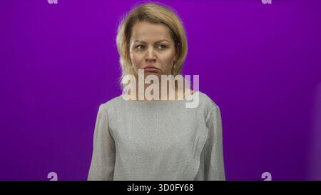 Young blonde woman pouting lips in purple studio setting wearing gray blouse, facing camera with a sullen expression; disappointment. Stock Photo