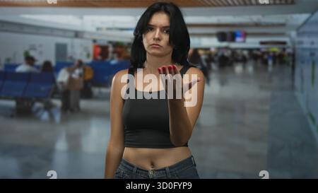 Young hispanic woman beckons with open hand at crowded airport terminal; delayed flight impatience. Stock Photo