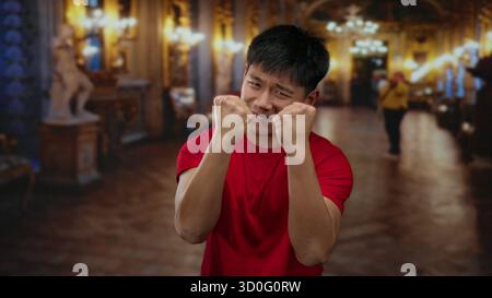 Young man in a red shirt poses with a determined expression in a richly decorated museum interior, showcasing a blend of historic art and modern emoti Stock Photo