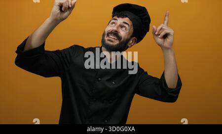 Hispanic male chef with a beard in black uniform joyfully pointing upwards against an orange background. Stock Photo