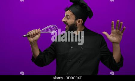 Hispanic chef with beard playfully sings using a whisk as a microphone against a vibrant purple background, exuding fun and creativity in culinary art Stock Photo