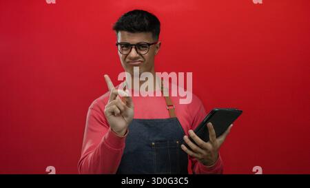 Young man wearing glasses and apron interacts with tablet against vibrant red background showcasing modern technology and casual style in a captivatin Stock Photo