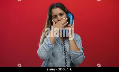 Woman holding blue telephone handset with hand over mouth laughing in red studio wearing light blue shirt and yellow nails; playful joy. Stock Photo