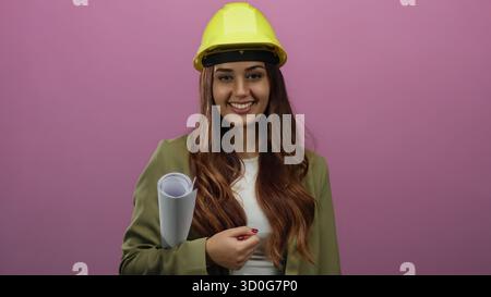 Young woman architect smiling confidently against an isolated pink background wearing safety helmet while holding rolled blueprints suggesting a profe Stock Photo