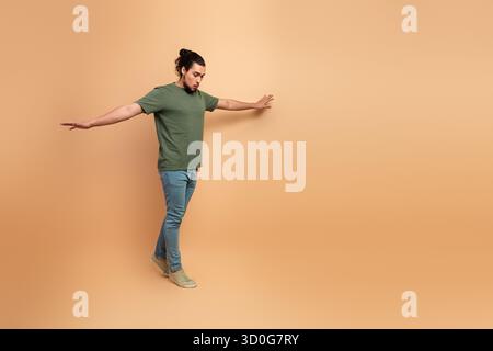 Young male model in green t shirt and jeans stands with arms outstretched against a beige background conveying casual fashion lifestyle Stock Photo